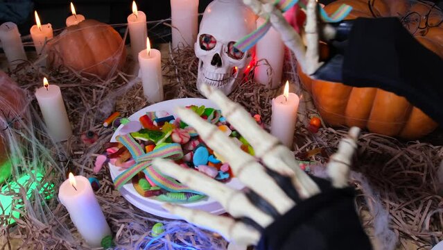 Creepy hand takes colorful candy sweets for trick or treat Halloween close up decor with flickering candles, skull and traditional pumpkins. Celebration Happy Halloween background.