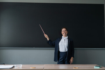 Red-haired caucasian woman in a trouser suit. Smiling female teacher with a pointer at the blackboard. 