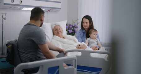Elderly woman lying and resting in bed in bright hospital ward, plays with grandson. Loving family...