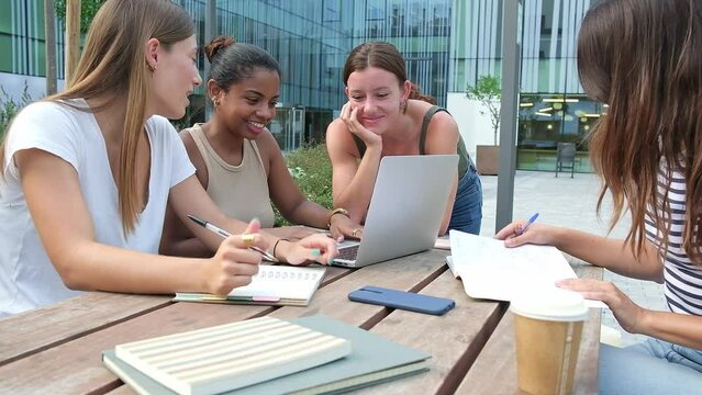 Multiracial female students studying and doing homework using laptops, laughing, talking outdoor in college campus.