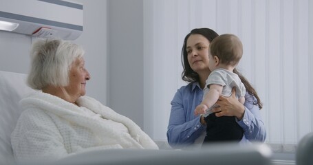 Fototapeta premium Elderly woman lying and resting in bed in bright hospital ward, plays with grandson. Loving family members support grandmother recovering after successful surgery. Modern medical facility or clinic.