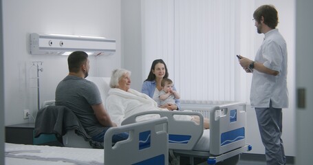 Female old patient lies in bed in hospital room, spends time with family. Male nurse, doctor uses digital tablet, consults elderly woman recovering after successful surgery. Modern medical facility.