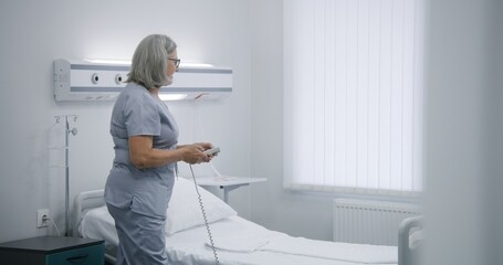 Mature nurse sets up bed using remote control, prepares hospital room for patient. Female medical worker pushes wheelchair with elderly woman and puts her in bed. Modern clinic or medical facility.