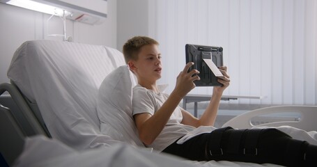 Young boy with bandaged leg lies on bed, talks with dad using tablet. Kid remotely shows hospital conditions to father. Patient fully recovering after successful surgery. Modern and bright clinic.