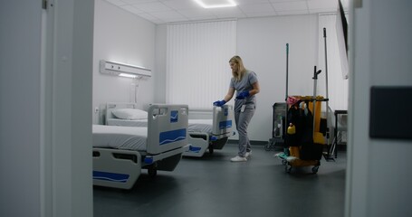 Adult nurse mops floor between beds in hospital ward. Health worker brings cleaning trolley to...