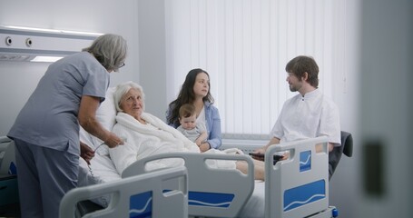 Elderly woman and her family in hospital room. Doctor uses digital tablet, consults female senior patient recovering after successful surgery. Nurse adjusts pillow and bed. Modern medical facility.