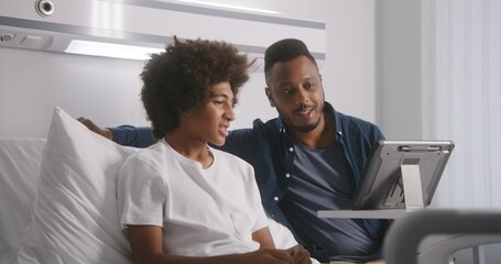 African American teenager rests on bed in hospital ward. Man visits son after surgery. Family...