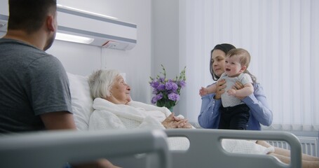 Fototapeta premium Female elderly patient lies in bed in bright hospital ward, talks to daughter with grandson. Loving family supports senior woman recovering after successful surgery. Modern medical facility or clinic.