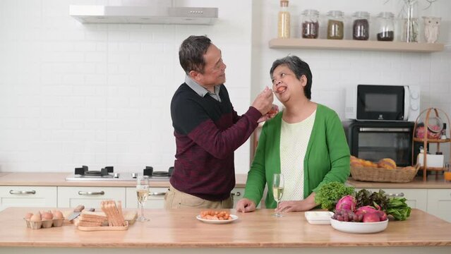 Happy Asian senior husband feeding sausage to his wife while standing at counter in kitchen