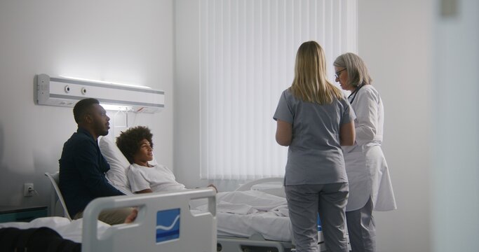 African American teen patient lies on bed in hospital room after surgery. Mature female doctor and nurse talk with teenager and father about his health. Medical personnel at work in modern clinic.