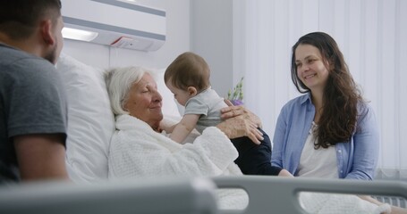 Obraz premium Female elderly patient lies in bed in bright hospital ward, plays with little grandson. Loving family members support senior woman recovering after successful surgery. Modern medical facility.