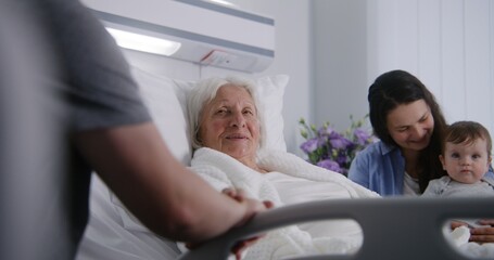 Elderly woman lying and resting in bed in bright hospital ward, plays with grandson. Loving family...
