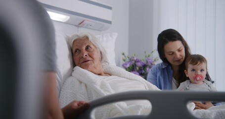 Elderly woman lying and resting in bed in bright hospital ward, plays with grandson. Loving family...