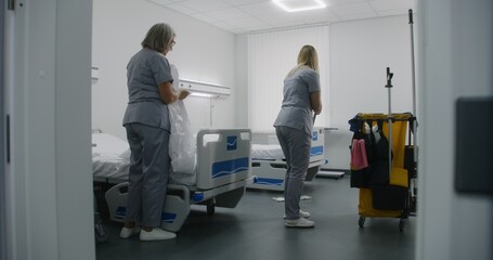 Obraz premium Health worker brings cleaning trolley to hospital ward. Nurses clean hospital room after patients. Mature cleaner with her colleague changes bedding on beds. Medical staff at work in modern clinic.