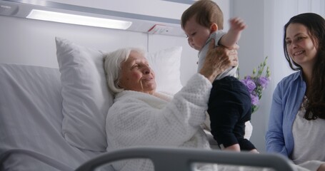 Female elderly patient lies in bed in bright hospital ward, plays with little grandson. Loving family members support senior woman recovering after successful surgery. Modern medical facility.