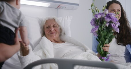 Obraz premium Loving family with little child visits grandmother recovering after successful surgery, gives flowers. Elderly woman lies and rests in bed in bright hospital ward. Modern medical facility or clinic.