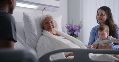 Female old patient lies in bed in bright hospital ward, spends time with relatives. Loving family...