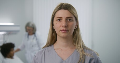 Obraz premium Adult nurse stands in hospital room with digital tablet computer in hands, looks at camera. Mature female doctor checks condition of patient in the background. Medical staff in modern medical center.