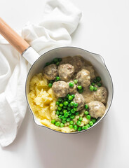 Delicious homemade lunch - mashed potatoes, Swedish meatballs in cream sauce and green peas in a saucepan on a light background, top view