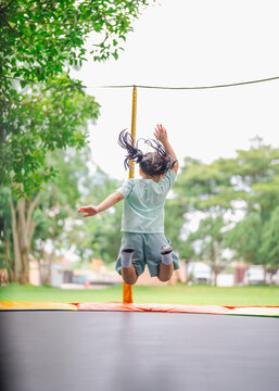 Asian child girl is jumping on trampoline on playground background. Happy laughing kid outdoors in the yard on summer vacation. Jump high on trampoline. Activity children in the kindergarten school.