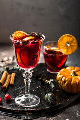 Glass of red wine with spices and fruits on a tray, in the background a glass of wine and orange, a small decorative pumpkin, cinnamon sticks, star anise and raspberries, in a dark style.