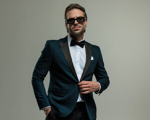 elegant young man in tuxedo holding hand in pocket and unbuttoning tux