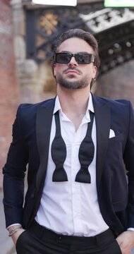 attractive elegant man with sunglasses holding hands in pockets, looking to side and adjusting suit and thinking 