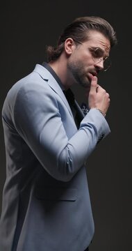 cool fashion guy with glasses looking down and moving, touching lips in a seductive manner, rubbing palms and holding hands in pockets on grey background