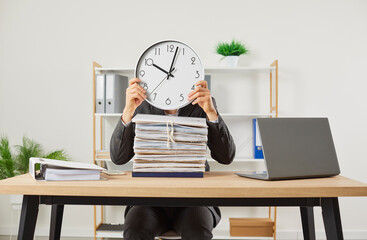 Overworked funny business man hiding head behind the clock at his workplace with a pile of folders...