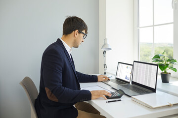 Male employee working in office. Financial accountant working with business data. Serious busy young man in suit and glasses sitting at desk by window and using calculator and two laptop computers