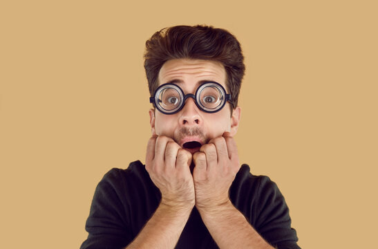 Stunned frightened young man in glasses with thick lenses. Portrait of a shocked guy covering mouth with hands, being surprised and scared to hear shocking news over isolated background