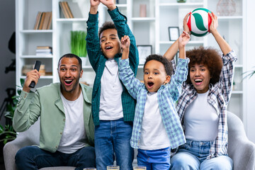 Joyful family raising arms in triumph and celebrating incredible victory. Father, mother and two sons watching volleyball game on tv sitting on couch at home