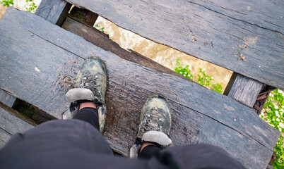 Feet in dirty trekking boots stand on a wooden suspension bridge over a stormy mountain river.
