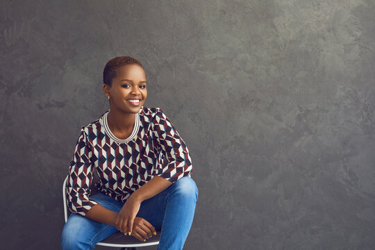 Studio Portrait Of Happy And Confident Dark Skinned Casual Woman Who Is Smiling While Looking At Camera. Woman In Casual Clothes Sitting On Chair On Gray Background. Place For Text. Advertising Banner