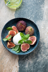 Blue bowl with fresh fig fruits, mozzarella and green basil, top view on a grey and beige stone background, vertical shot