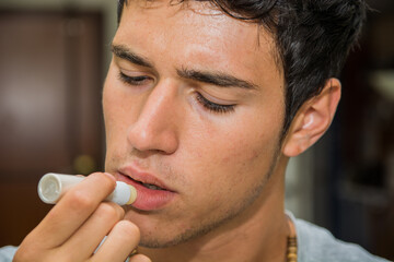 A man with a necklace on putting a li balm on his mouth. Photo of a young man moisturizing his lips with a lip balm stick - obrazy, fototapety, plakaty