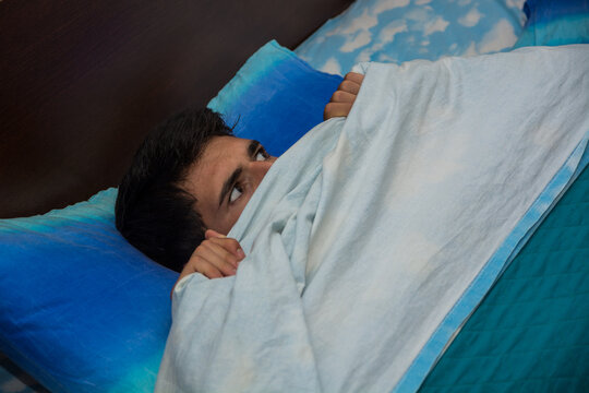 A person laying in a bed with a blue pillow, with his eyes wide open, scared from something he heard in the house