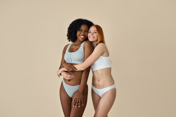 Two happy diverse women, African and Caucasian young girls friends multiethnic models hugging smiling looking at camera standing isolated on beige background. Diversity and body beauty concept.