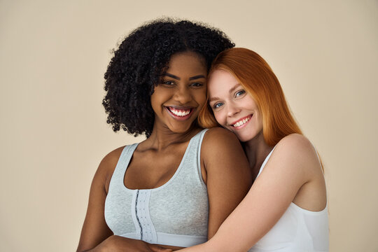 Two happy diverse women, African and Caucasian young girls friends multiethnic models hugging smiling looking at camera standing isolated on beige background. Diversity and beauty concept, portrait.