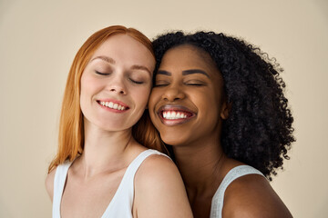 Two smiling diverse cute young girls bonding eyes closed isolated on beige background. Happy...