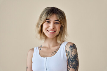 Young happy blond pretty smiling girl beauty female gen z model with short blonde hair beautiful face healthy skin and tattoo looking at camera wearing white top isolated at beige background. Portrait