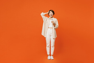 Full body young shocked happy caucasian woman she wear beige shirt casual clothes hold head use mobile cell phone look camera isolated on plain orange red background studio portrait Lifestyle concept