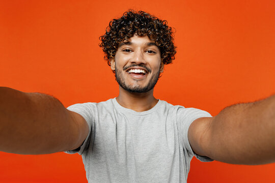 Close up young smiling satisfied happy Indian man he wears t-shirt casual clothes doing selfie shot pov on mobile cell phone isolated on orange red color background studio portrait. Lifestyle concept.