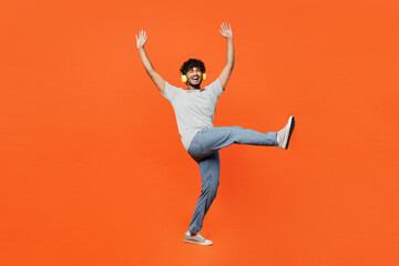 Fototapeta na wymiar Full body young smiling happy Indian man he wears t-shirt casual clothes listen to music in headphones raise up hands dance isolated on orange red color background studio portrait. Lifestyle concept.