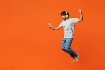 Fototapeta na wymiar Full body side view young Indian man he wearing t-shirt casual clothes jump high use mobile cell phone listen to music in headphones isolated on orange red color background studio. Lifestyle concept.