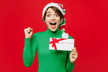 Young excited woman wear green turtleneck Santa hat posing hold gift certificate coupon voucher...