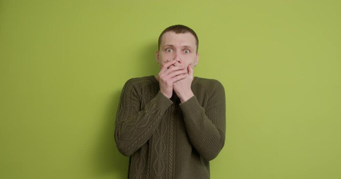 Young scared emotional man covering mouth with hands. Can not speak over isolated green background. Slow motion, nervous guy looking through fingers, palms, touching head, does not know what to do