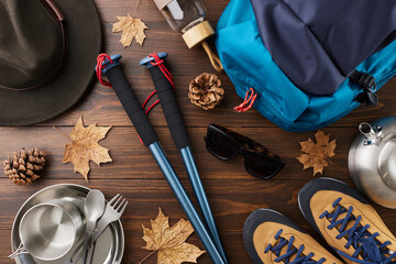 Autumn adventure tourism concept. Top view shot of backpack, eyewear, hat, trekking sticks, boots, metal utensils, bottle, dry autumn leaves, cones on wooden background with promo spot