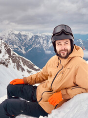 A handsome young man in ski gear lies on the snow on a mountain slope and smiles. A snowboarder at a ski resort