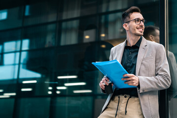 Smiling businessman holding paper documents and takeaway coffee outdoors. Confident young man with...